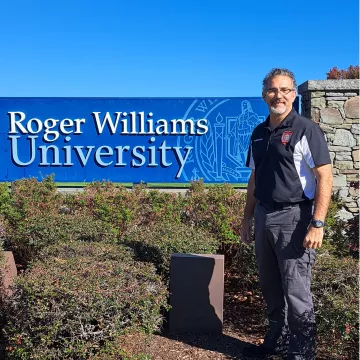 John Scott standing in front of the Roger Williams University sign after delivering a talk to Criminal Justice students at RWU.