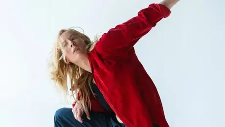 Woman dancing with foot and arm in the air while wearing red sweater and jeans.