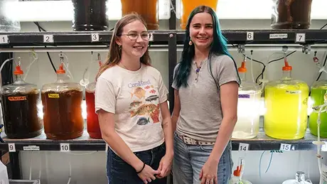 Cara Harvey and Gillian Frey stand near carboys of algae.