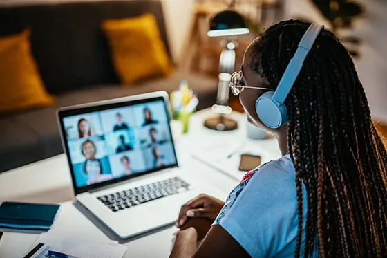 A student sits looking at her laptop wearing headphones and participating in a virtual meeting