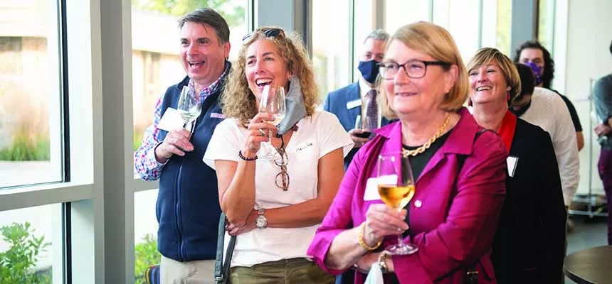 Three alumni stand smiling and holding wine glasses at the front of a well-attended alumni event