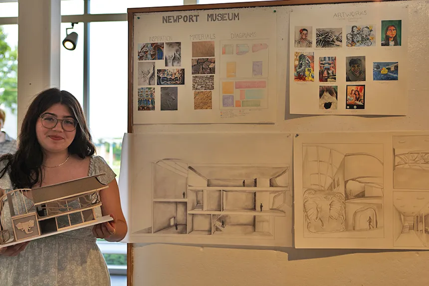 A student standing with her model and her work pinned on a gallery wall 