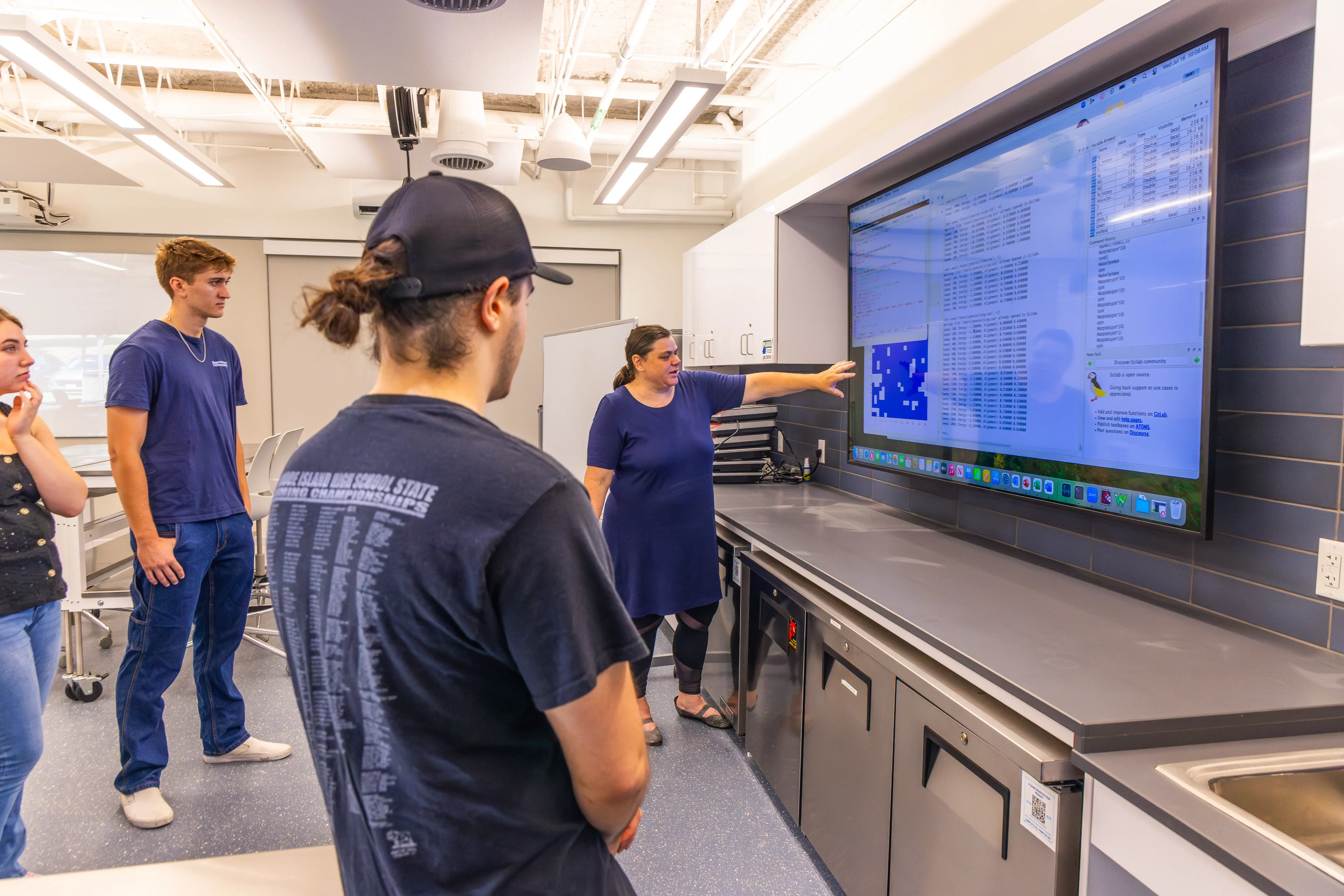Instructor points at data on a large screen while students watch attentively in a modern classroom lab.