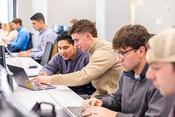 6 students pictured sitting at a table looking at laptops. 
